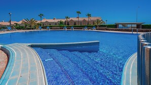 En klarblå pool med ett kaklat poolområde, omgiven av solstolar och palmer, och en byggnad i bakgrunden.