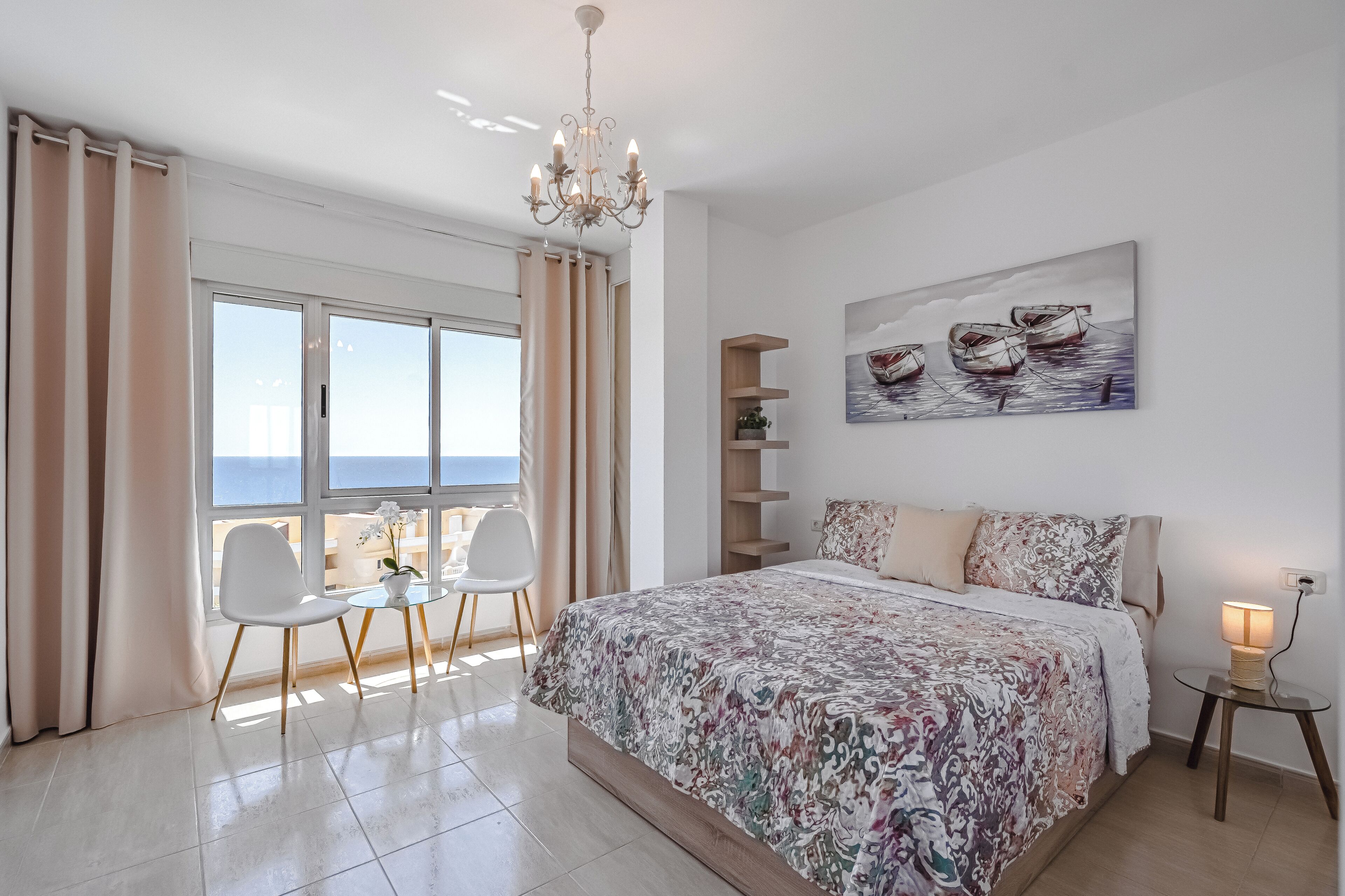Een slaapkamer met een groot bed, een kroonluchter, een schilderij van boten, een klein tafeltje met een lamp en uitzicht op de zee.