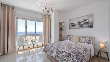 Een slaapkamer met een groot bed, een kroonluchter, een schilderij van boten, een klein tafeltje met een lamp en uitzicht op de zee.