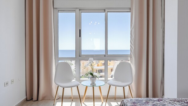 Ett rum med en säng, två vita stolar, ett litet glasbord med en blomma och ett stort fönster med utsikt över havet och byggnaderna.
