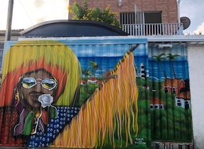 A colorful mural on a building's exterior wall, depicting a figure with a long, flowing garment and a tropical background with palm trees and houses. - A Joia de Olinda (Olinda)
