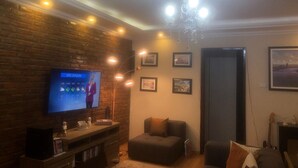 Een woonkamer met een bakstenen muur, een televisie waarop een weersvoorspelling te zien is, een kroonluchter, ingelijste foto's en een bank.