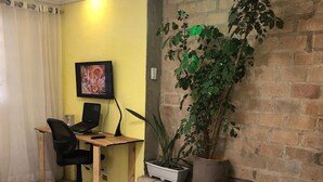 Een kamer met een houten bureau, een stoel, een laptop, een televisie, een potplant, een dienblad met een kop en een vaas, en een bakstenen muur.