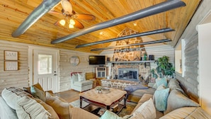 A cozy cabin interior with a stone fireplace, wooden ceiling beams, and a comfortable seating area.