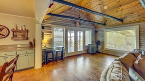 A spacious living area with a wooden ceiling, a kitchen with light-colored cabinets, and a large window.