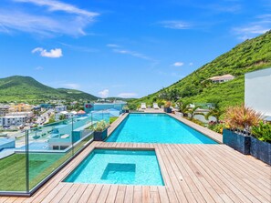 A rooftop pool with a wooden deck, surrounded by lush greenery and a view of a town and hills in the distance. - A132 - Glorious Lagoon View Apartment with Balcony (Sint Maarten)