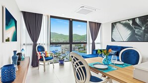 A modern dining area with a view of the sea and hills, featuring a wooden table, blue chairs, and a blue sofa. - A133 -  Top Floor Lagoon View (Sint Maarten)