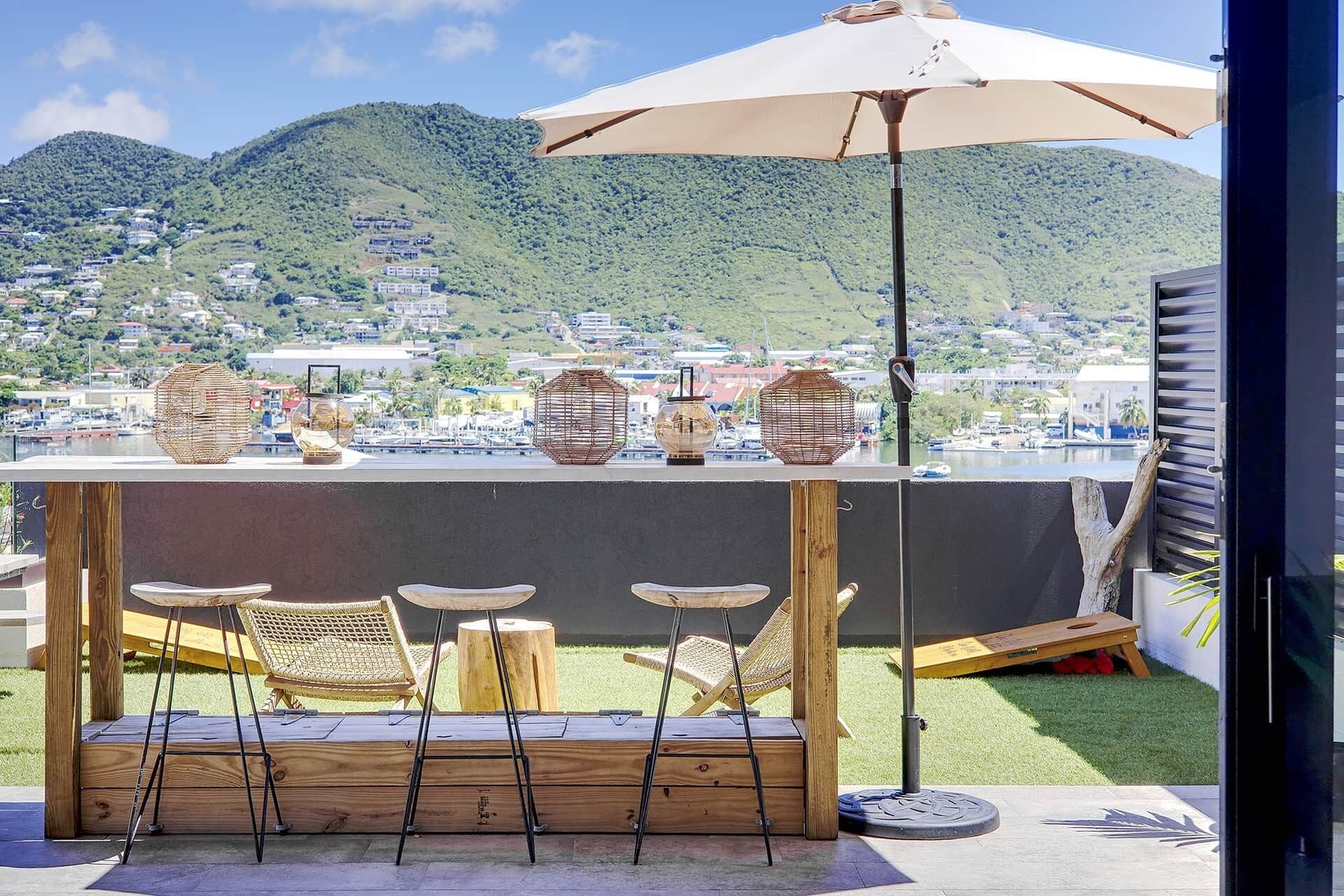 Una terraza con bar, sillas de mimbre y vistas a un puerto deportivo y colinas.