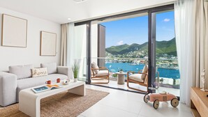 Una sala de estar moderna con sofá, mesa de centro y sillones, con vistas a un puerto deportivo y montañas a través de amplios ventanales.