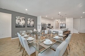 A modern dining area with a glass table, white chairs, and a kitchen in the background. - Amazing 15 Suites Mansion/Pool/Spa/Close to Disney (Kissimmee)