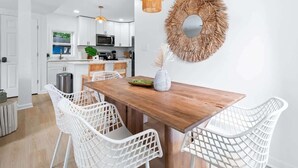 Un comedor con una mesa de madera y sillas de mimbre blanco, un adorno de pared tejido y una cocina al fondo.