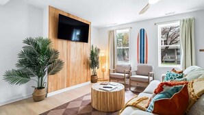 Una sala moderna con un televisor de pantalla plana montado en una pared de madera, un ventilador de techo, un ventanal con cortinas, un sofá con cojines de colores y dos plantas en macetas.