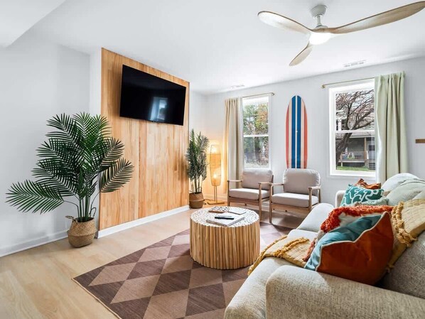 A modern living room with a flat-screen TV mounted on a wooden wall, a ceiling fan, a large window with curtains, a sofa with colorful cushions, and two potted plants. - 5 Min to Cedar Point Hot Tub Retreat (Sandusky)