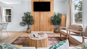 Una sala de estar moderna con un televisor de pantalla plana montado en una pared de madera, dos plantas en macetas, una mesa de centro redonda con un juego de mesa y un sofá con cojines.
