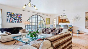 A spacious living room with a large window, a dining area, and a kitchen in the background.