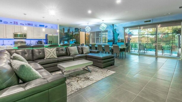 A modern living room with a sectional sofa, a kitchen area, and a dining space with a view of the cityscape. - Brand New 15BR Mansion/Pool/Spa/Close to Disney (Kissimmee)