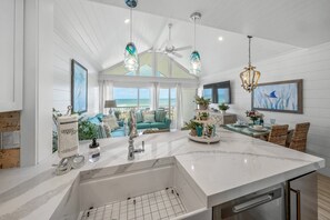 A spacious kitchen with a large island, a dining area with wicker chairs, and a view of the ocean through large windows.