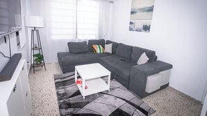 A modern living room with a grey sectional sofa, a white coffee table, a floor lamp, and a large window with sheer curtains. - 3BR Apartment w/ HDTV, 5 mins from Beach, pets OK.  (Río Grande)