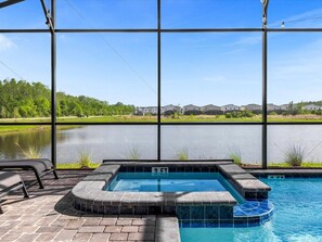 A swimming pool with a hot tub, surrounded by a brick patio and large windows offering a view of a lake and residential buildings. - Brand New Storey Lake 9BR Villa/Summer Kitchen (Kissimmee)