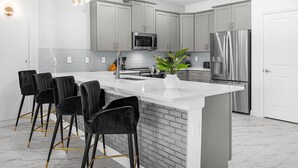 A modern kitchen with a large island, black bar stools, stainless steel appliances, and a white backsplash. - Brand New Storey Lake 9BR Villa/Summer Kitchen (Kissimmee)