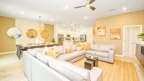 Ein modernes Wohnzimmer mit einem Ecksofa, einem Essbereich mit Tisch für vier Personen, einer Küche im Hintergrund und einem Deckenventilator.