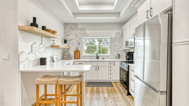 A modern kitchen with white cabinetry, a stainless steel refrigerator, wooden stools, and a window providing natural light.
