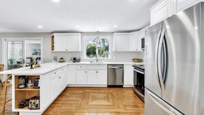 A modern kitchen with white cabinetry, a stainless steel refrigerator, and a wooden floor. - Luxury retreat, in-ground huge pool, 10-person hot tub, outdoor shower, fire pit (Barnstable)