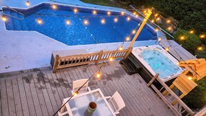 A hot tub with a wooden deck, string lights, and lounge chairs by a swimming pool. - Luxury retreat, in-ground huge pool, 10-person hot tub, outdoor shower, fire pit (Barnstable)