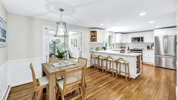 A modern kitchen with white cabinetry, stainless steel appliances, and a wooden dining area. - Luxury retreat, in-ground huge pool, 10-person hot tub, outdoor shower, fire pit (Barnstable)