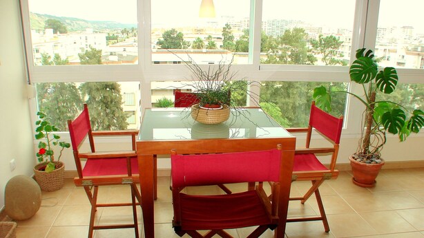 A dining area with a glass table and wooden chairs, a potted plant, and a view of a cityscape through large windows.