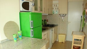 A compact kitchen with a green refrigerator, a microwave, and a small sink. There is a wooden stool and a small shelf with kitchen items.