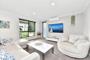 Ein modernes Wohnzimmer mit Sofa, Couchtisch, Fernseher und einem großen Fenster mit Blick auf Grünflächen.