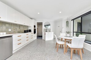 Een moderne keuken met witte kasten, een eethoek met een ronde tafel en stoelen, en uitzicht door een groot raam.