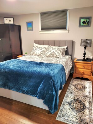 Ein Schlafzimmer mit einem Bett, einem Nachttisch, einer Lampe, einem Fenster mit Jalousien und einem Wandbild.