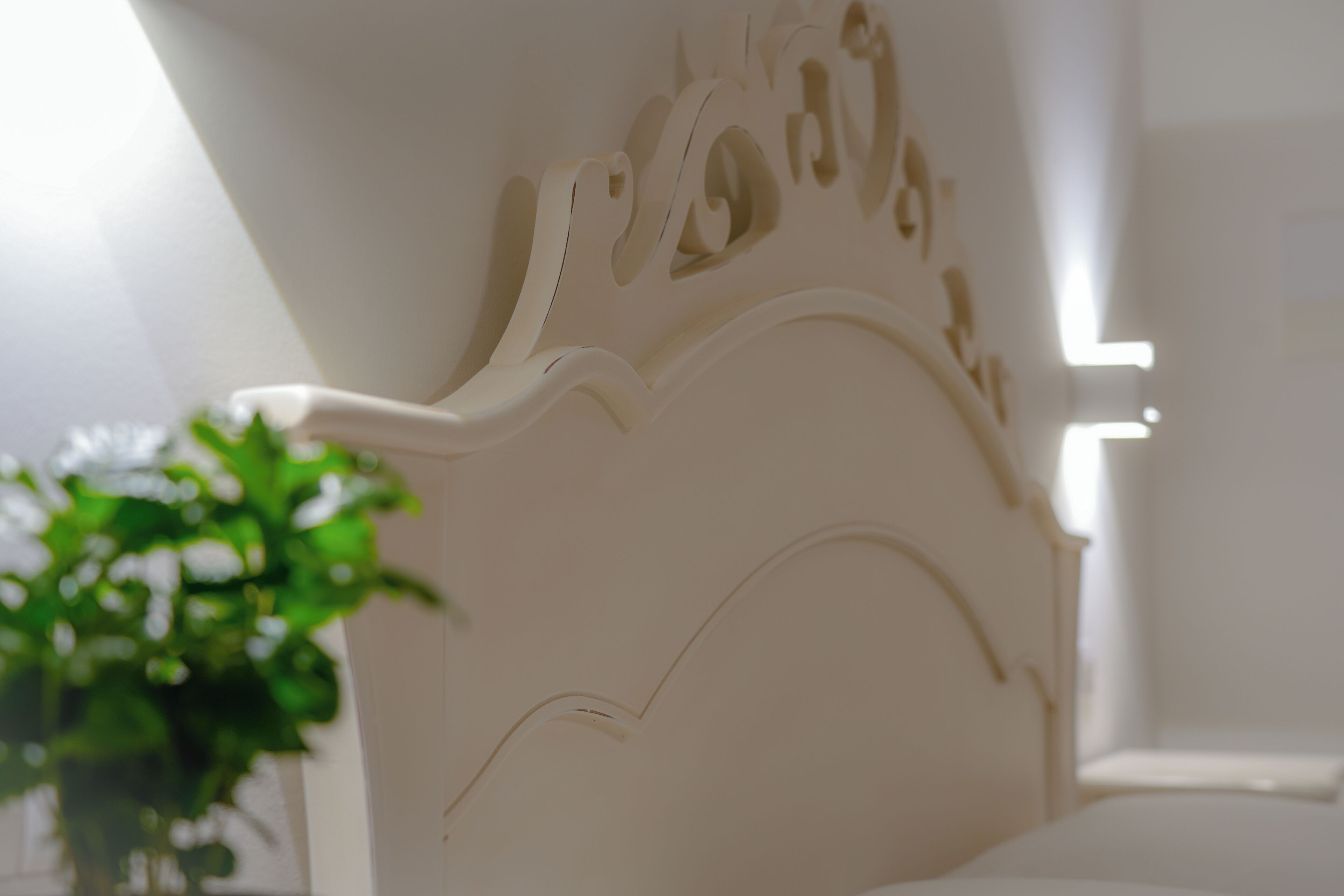 A white headboard with intricate carvings, a potted plant, and a window with natural light.