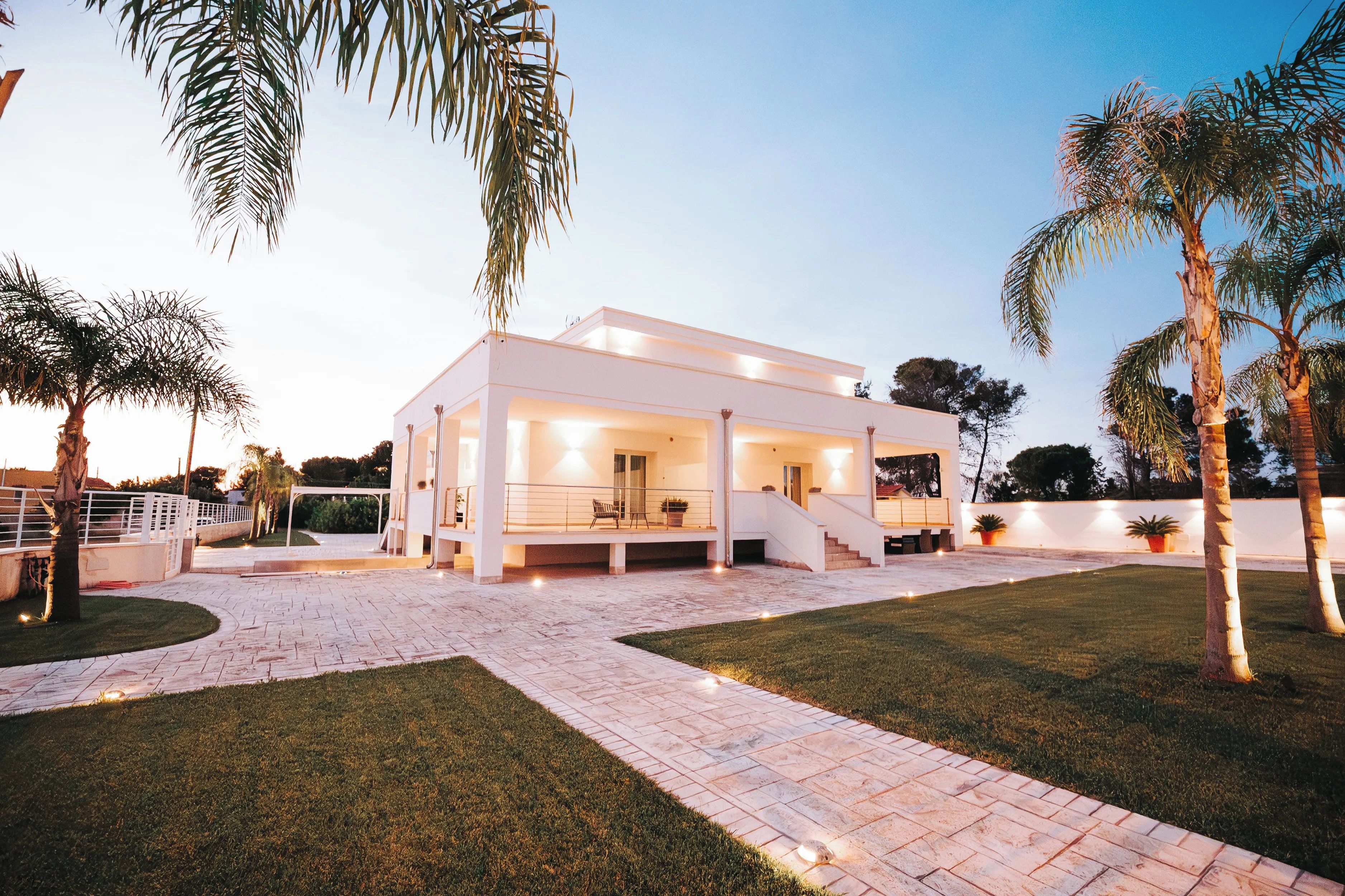A modern two-story house with a covered patio, a stone pathway, and palm trees.