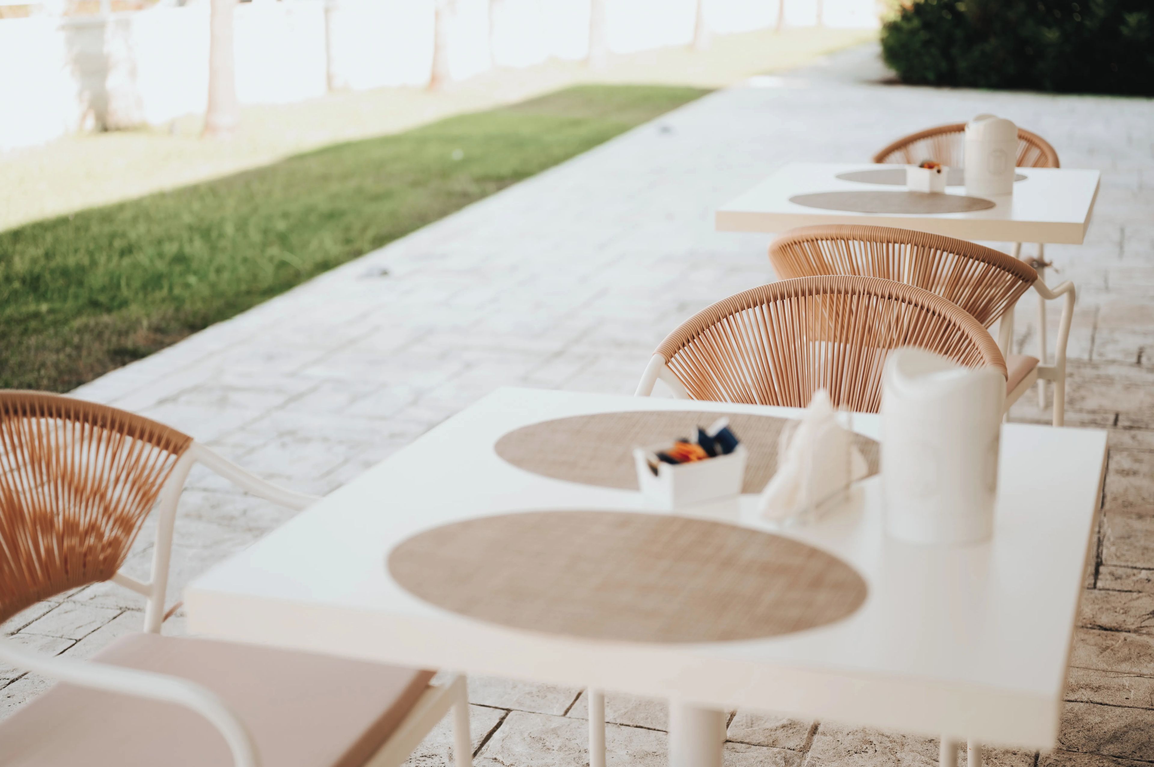 Outdoor seating with white tables and wicker chairs, arranged on a paved patio.