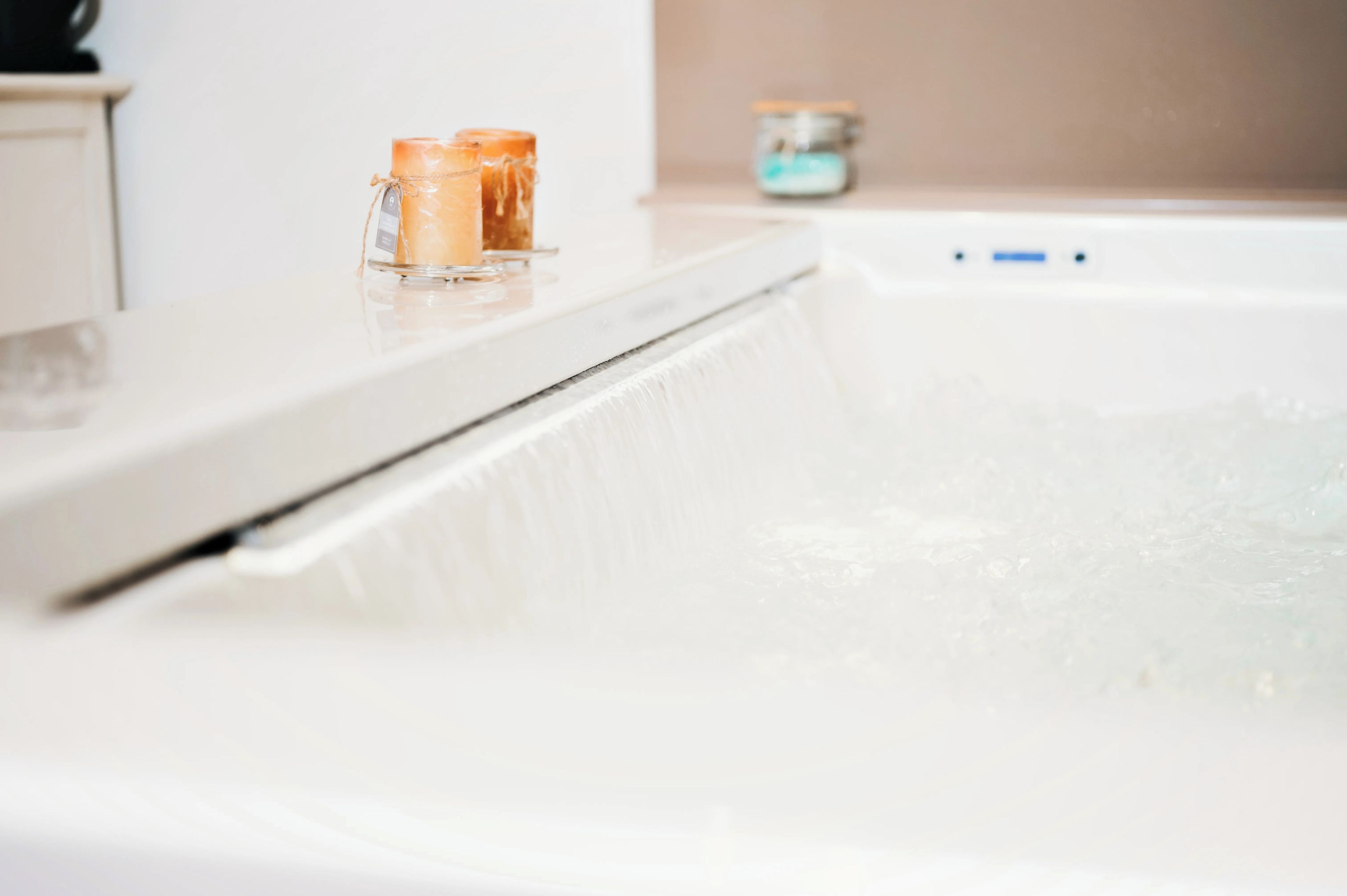 Una bañera con agua corriendo, un tarro de cristal y dos velas sobre una encimera blanca.