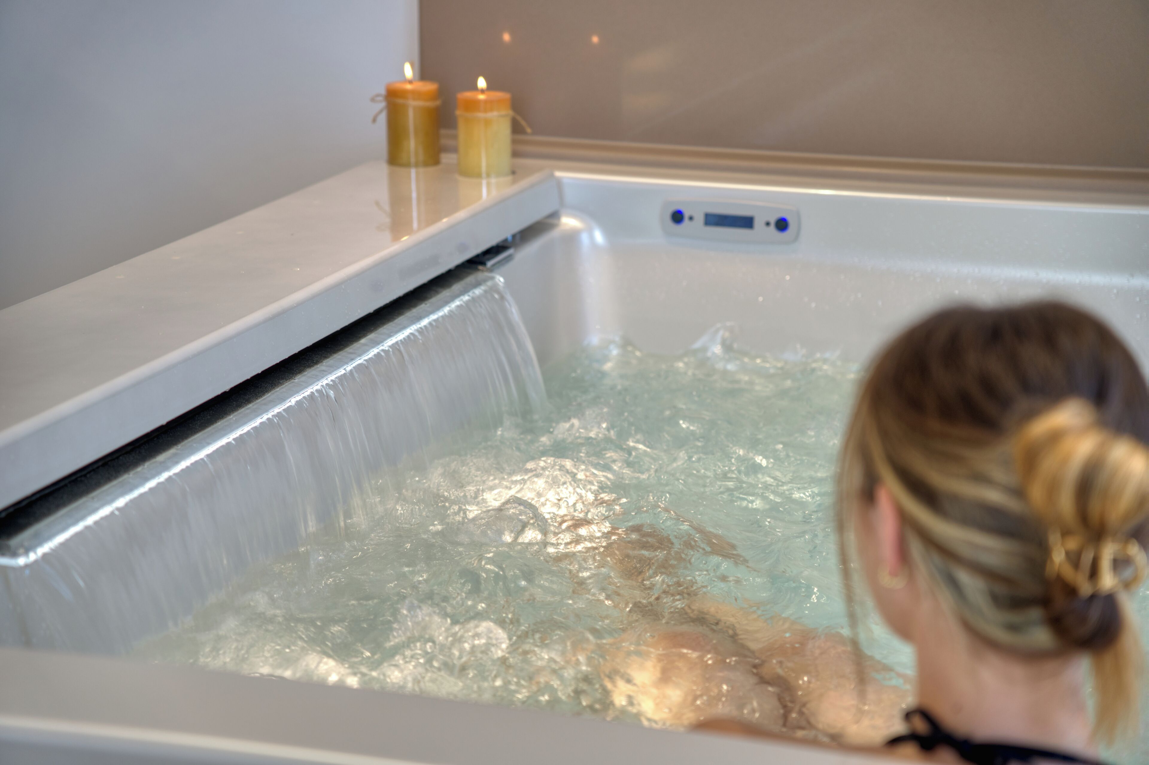 A person in a bathtub with a water feature, candles, and a control panel.