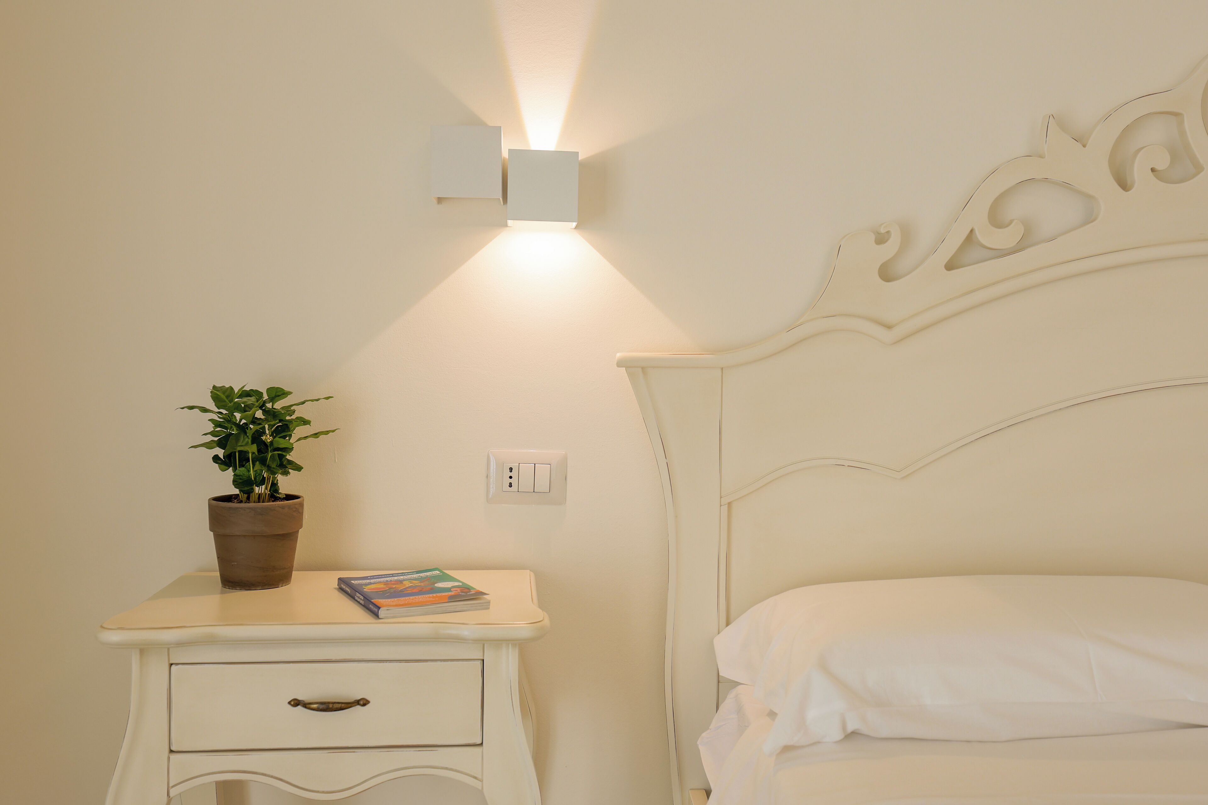 A bedroom with a white bed, a nightstand with a potted plant and a book, and a wall-mounted light fixture.