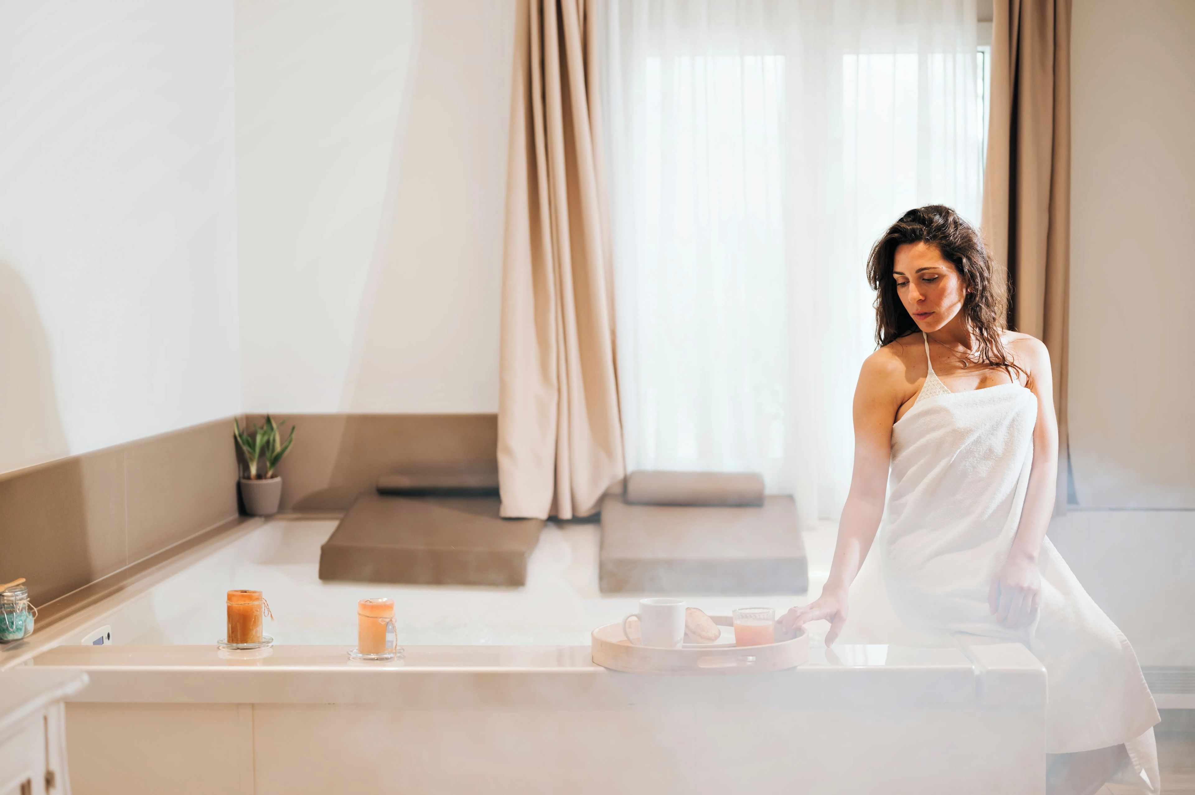 A person in a white towel standing by a counter with a tray containing a mug, a glass of orange juice, and a pastry.