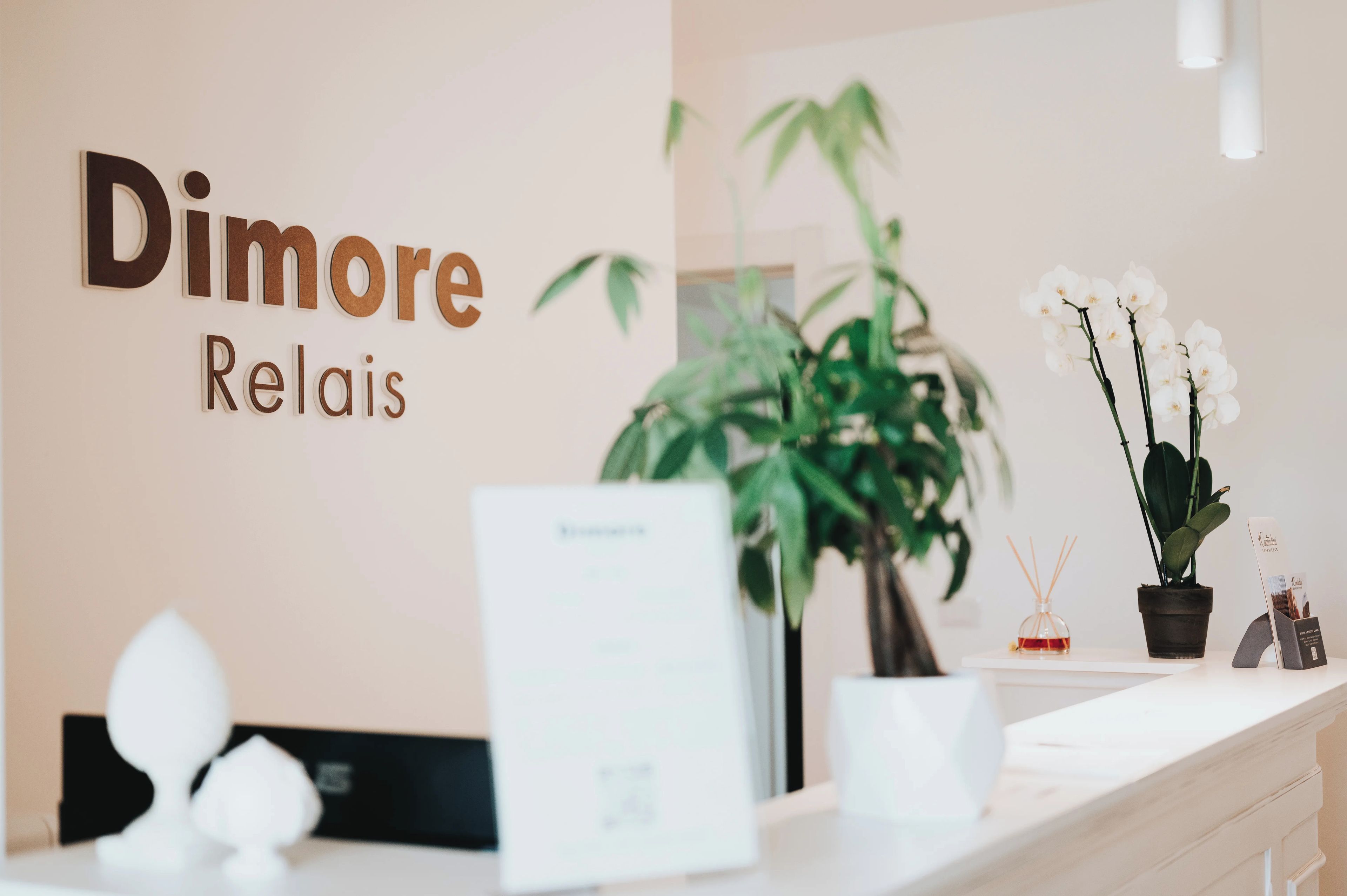 Een receptiebalie met een bordje met de tekst 'Dimore Relais'. Er staat een potplant en een display met witte orchideeën op de balie.