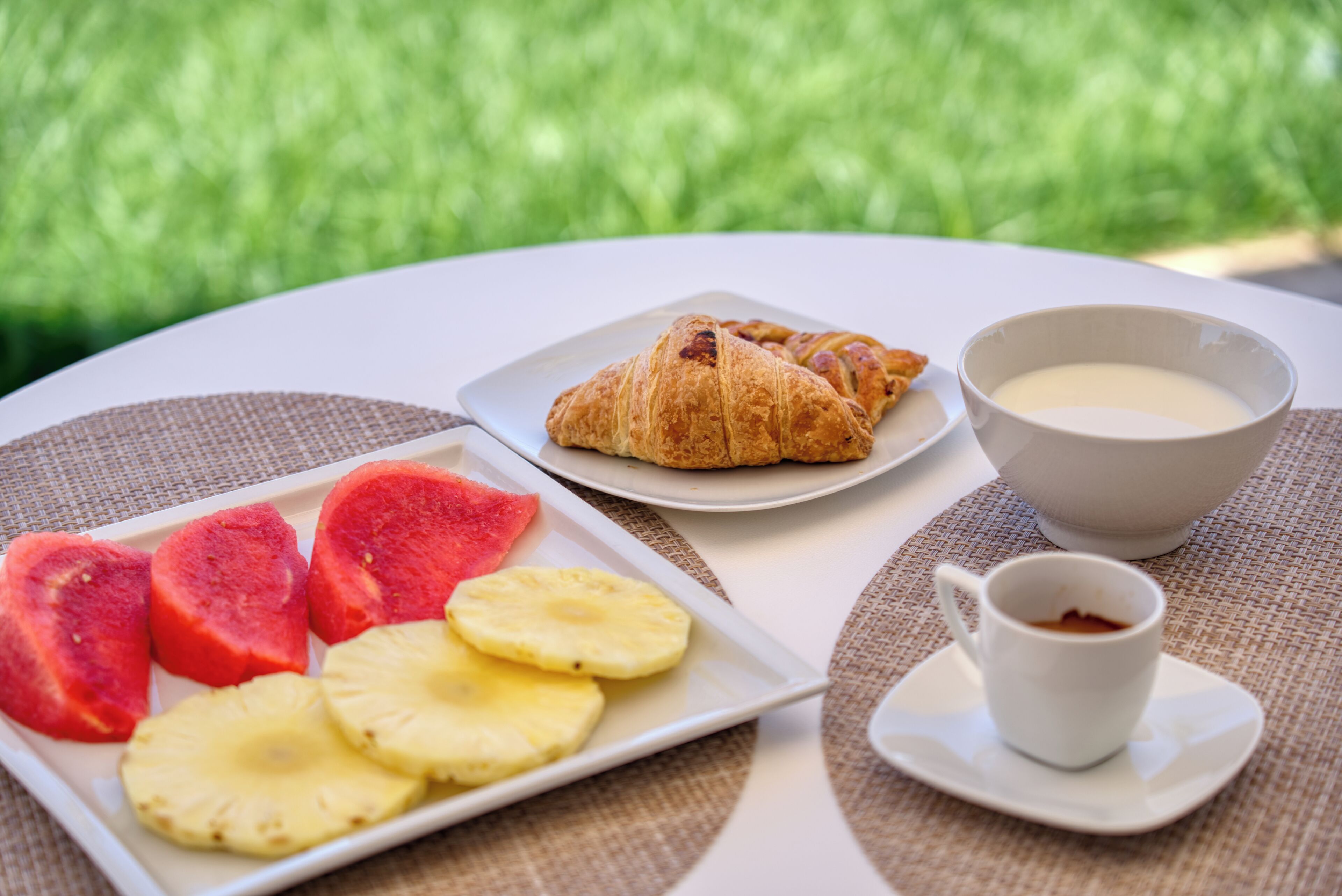 A breakfast setting with croissants, yogurt, coffee, and tropical fruit on a white table outdoors.