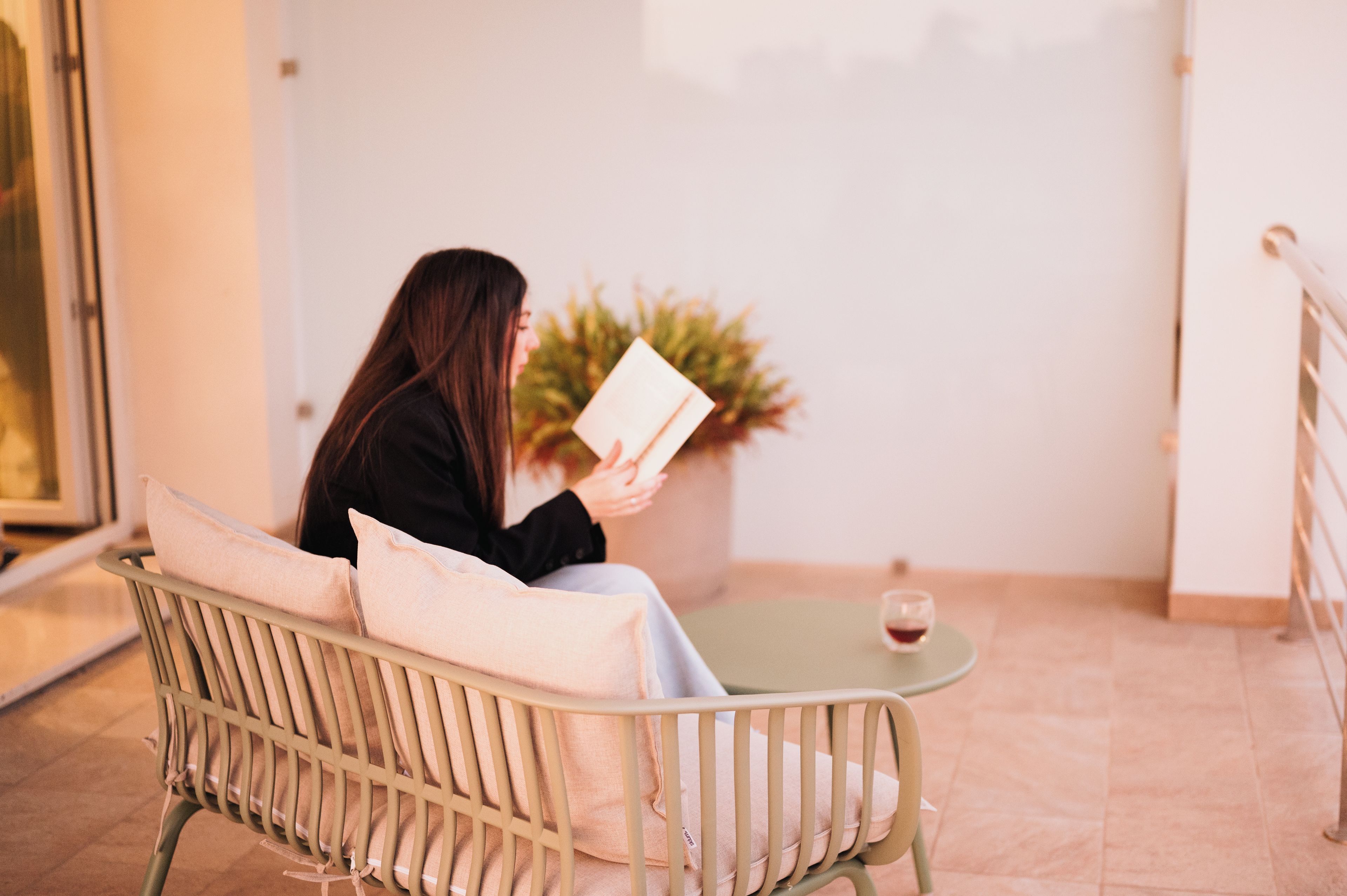 A person seated on a chair with a cushion, reading a book, with a glass of red wine on a small table beside them.