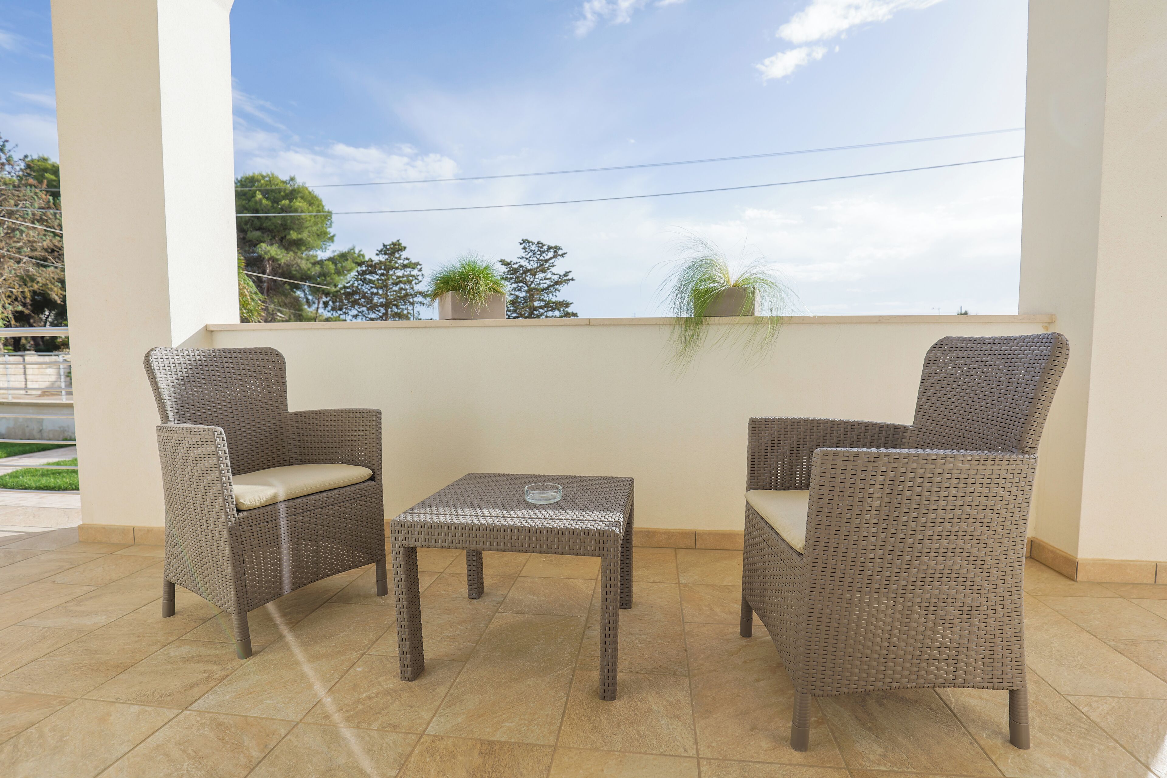 A balcony with wicker furniture, including two chairs and a small table, and potted plants on the railing.