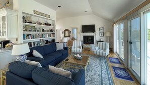 A spacious living room with a large sofa, a fireplace, and a bookshelf filled with books and decorative items. - Gorgeous South Beach Contemporary 5 min walk to beach with AC, and WiFi (Edgartown)