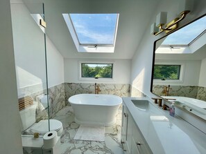 Un baño moderno con una bañera independiente, un espejo grande y un tragaluz sobre el inodoro.