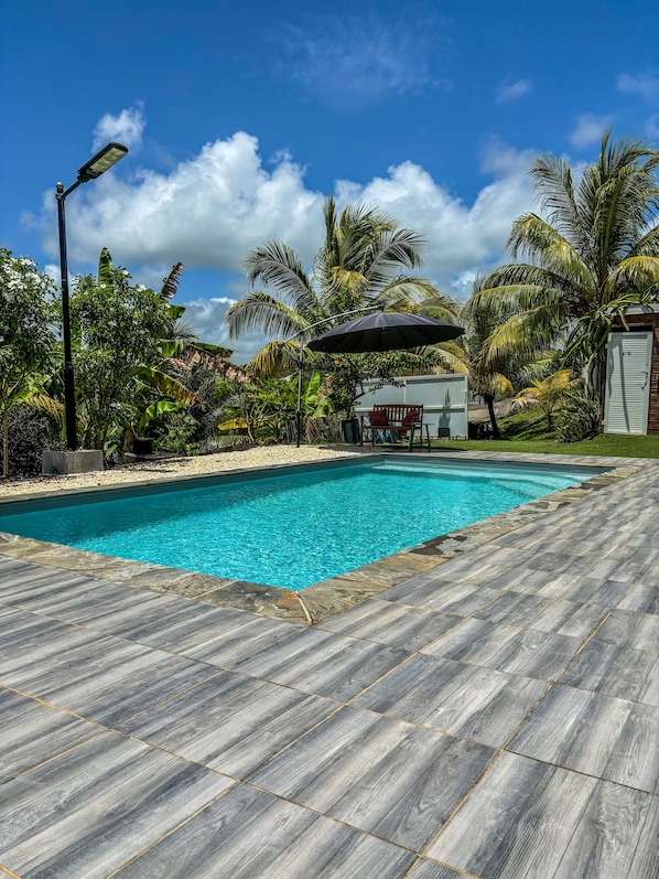 A swimming pool with a wooden deck, surrounded by tropical plants and palm trees.