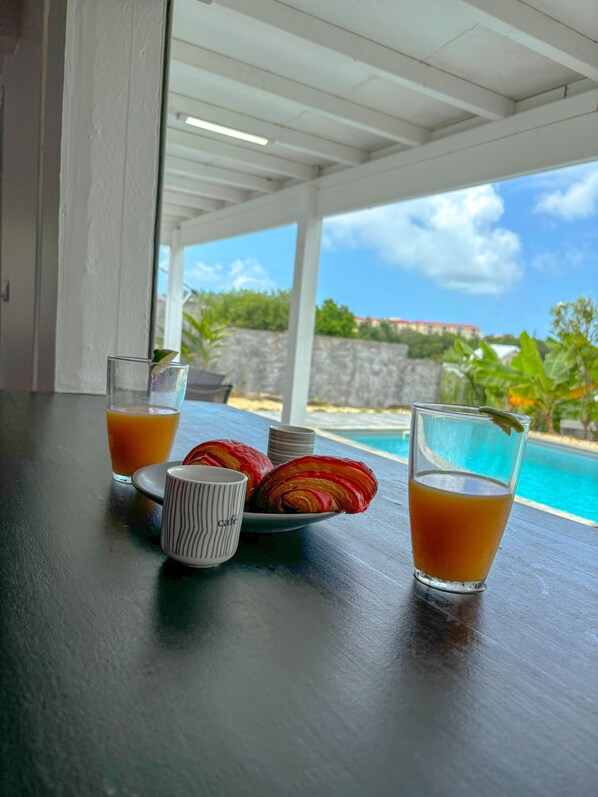 A table with croissants, a glass of orange juice, and a mug, overlooking a pool and greenery.
