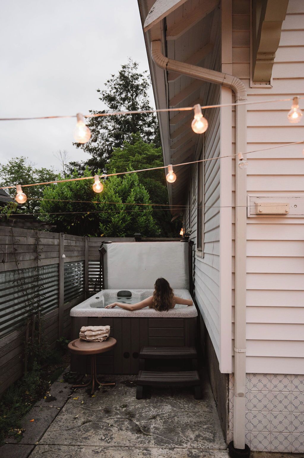 Un jacuzzi en un patio trasero con valla de madera, escalones e iluminación colgante.